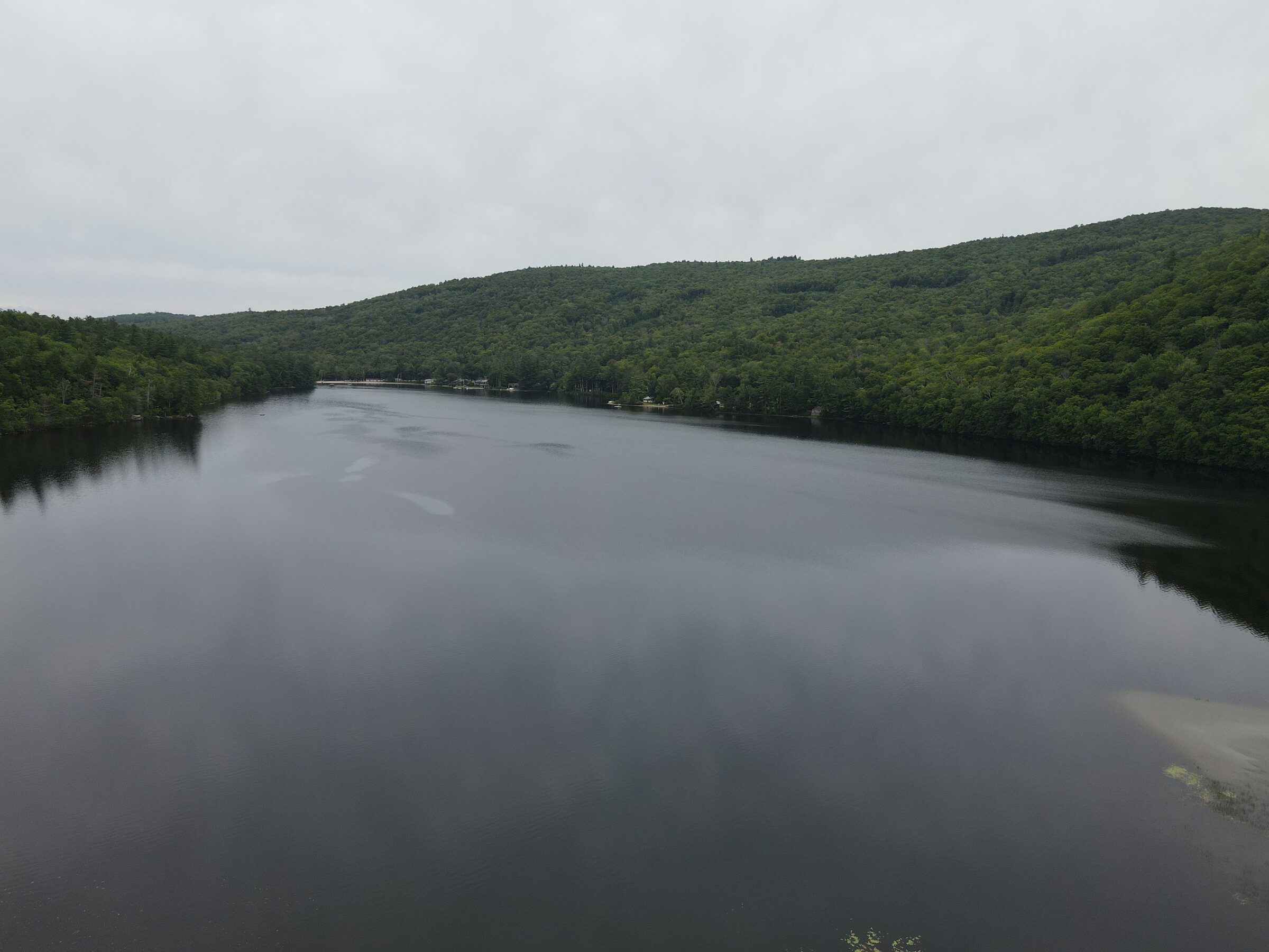 Aerial view of a still lake below wooded hills on an overcast day